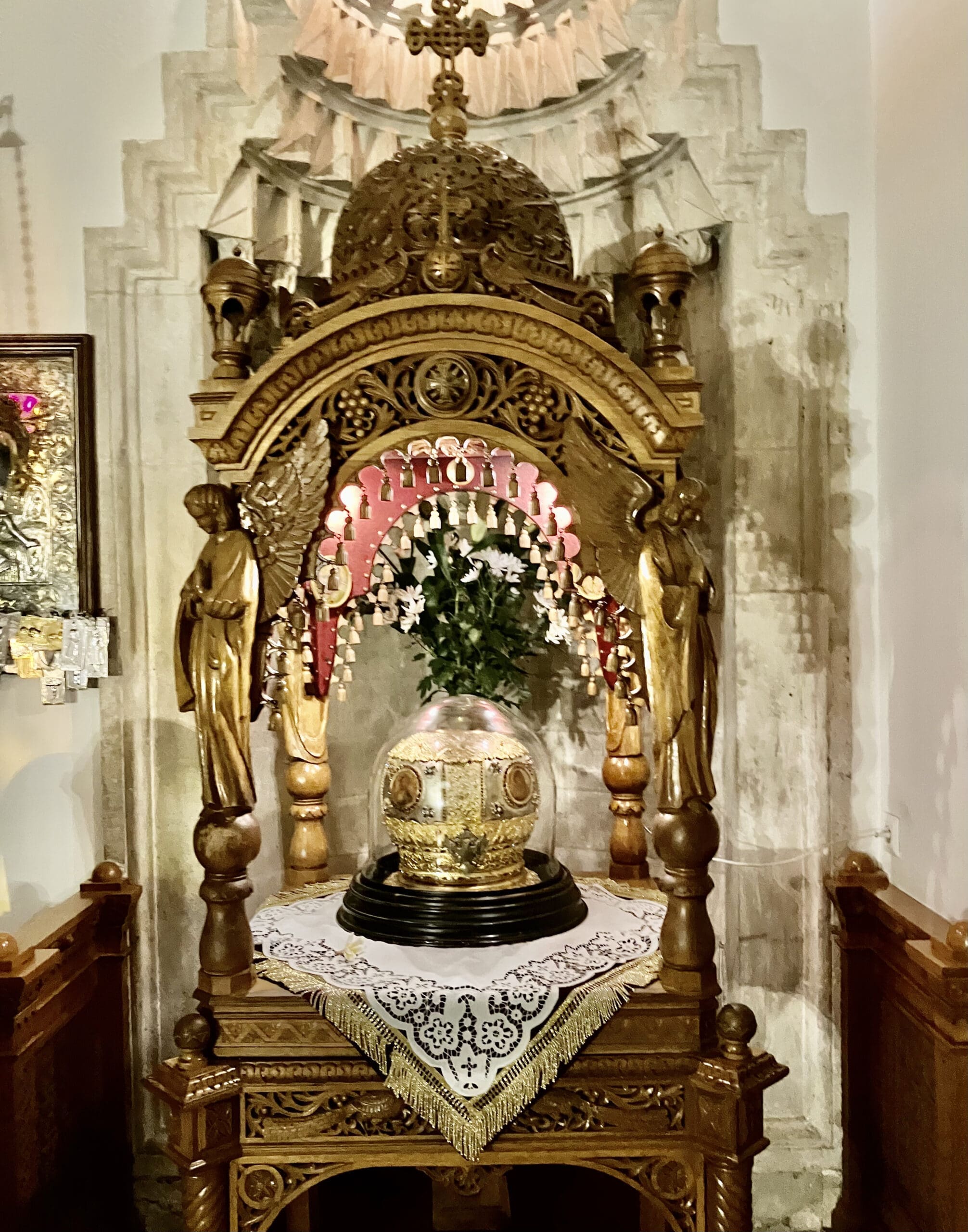 A golden religious relic or ornate crown is displayed under a decorative wooden canopy with carved figures, inside what appears to be a church or chapel. The relic sits on a lace-covered table.