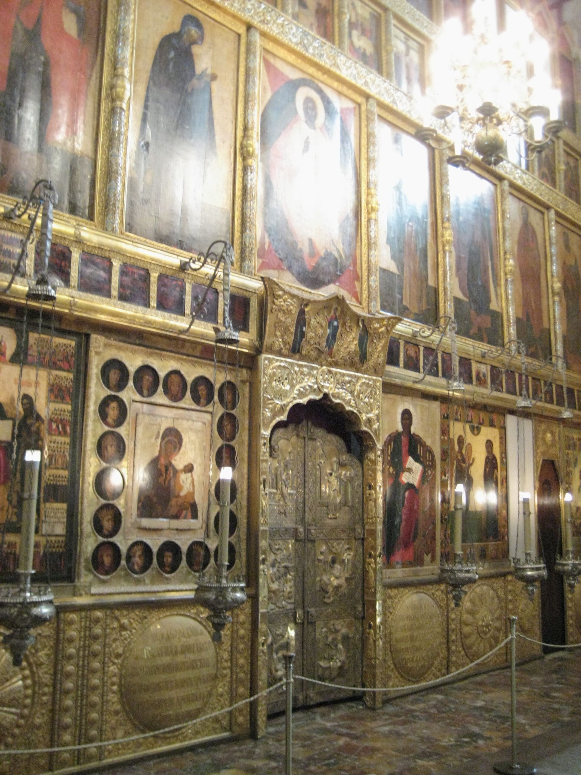 A richly decorated Russian Orthodox iconostasis with gold accents, religious icons, and ornate double doors, illuminated by a chandelier, inside a church. Rope barriers separate the iconostasis from visitors.