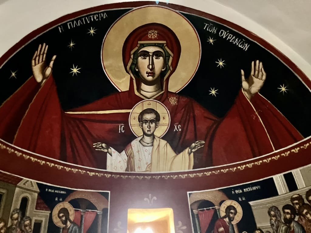 Religious mural showing the Virgin Mary with outstretched arms and the Christ Child in front of her, surrounded by a halo and Greek text, with gold stars on a dark background above other smaller religious figures.