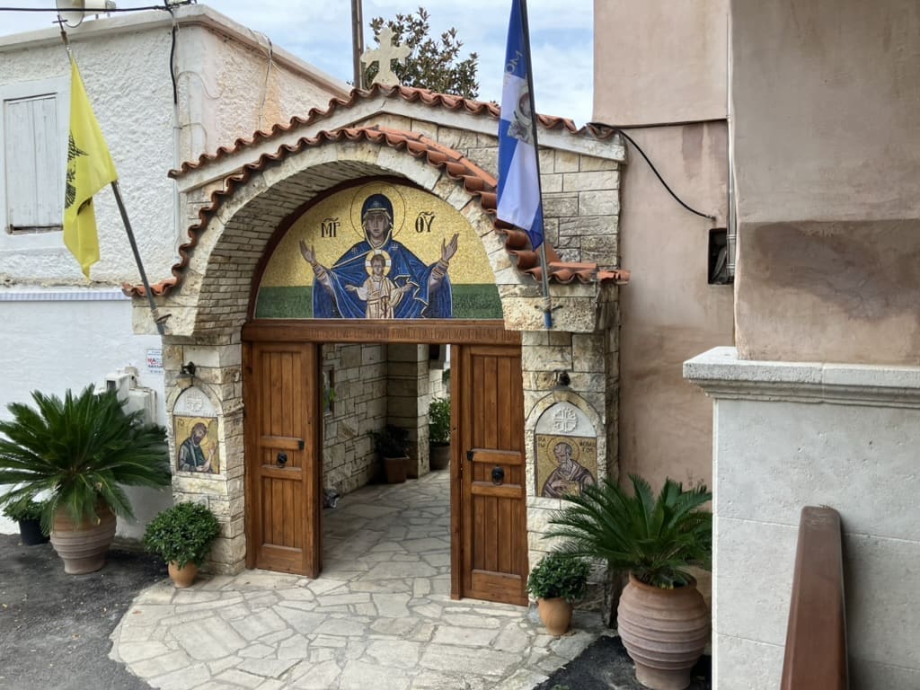 A stone entrance with wooden doors at a Greek Orthodox church, featuring a mosaic of the Virgin Mary and child above the archway, flanked by two icons and potted plants. Greek and religious flags hang above.