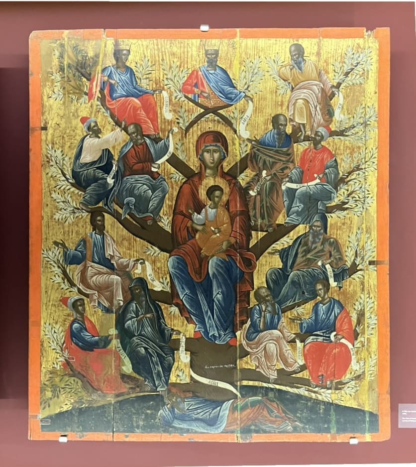 A religious icon painting shows a central female figure with a child, seated on a tree with multiple male figures in colorful robes sitting on its branches, set against a golden background.