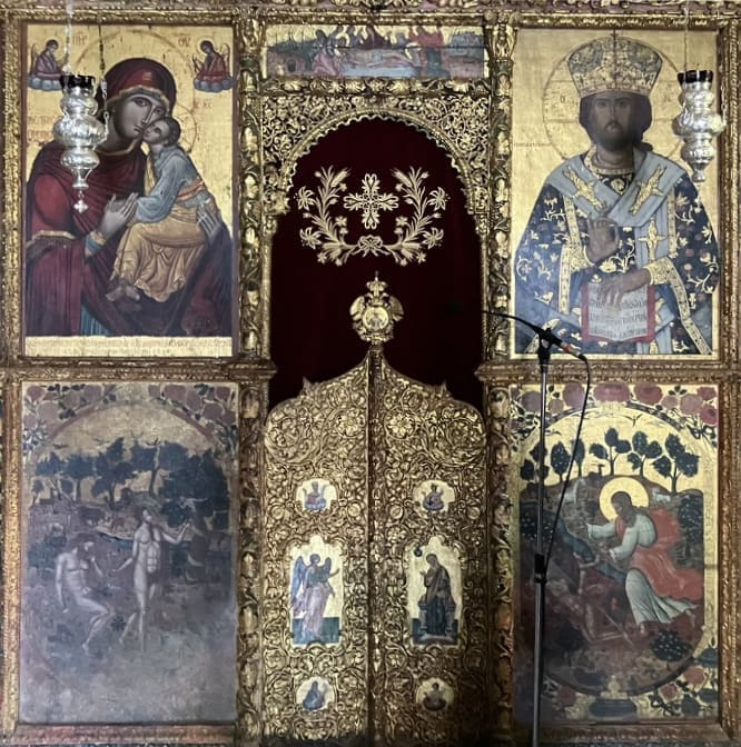 An ornate gilded church iconostasis featuring religious paintings of the Virgin Mary with Jesus, a crowned saint holding a staff, biblical scenes, and intricate gold detailing; microphones stand nearby.