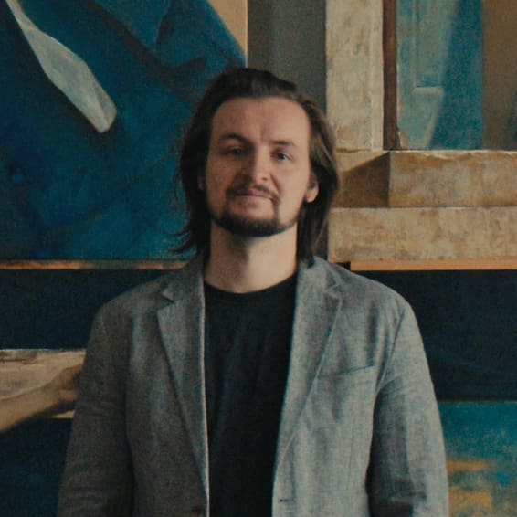 A person with medium-length brown hair and a beard, wearing a gray blazer over a black shirt, stands in front of a wall with blue and beige abstract artwork.