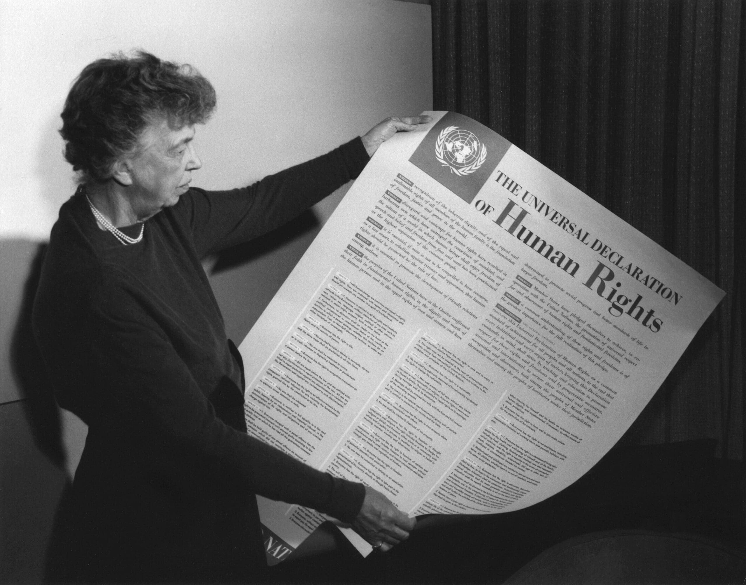 A woman stands holding and reading a large printed copy of the Universal Declaration of Human Rights, featuring the United Nations emblem at the top.