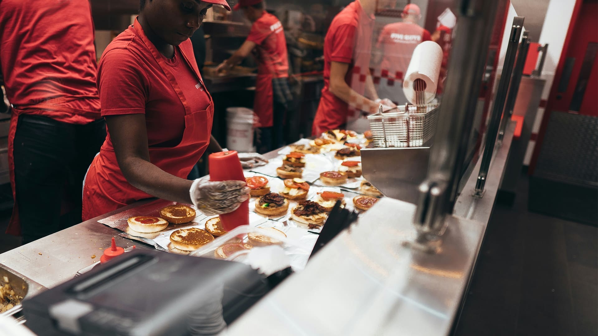 Fast food workers in red uniforms prepare multiple burgers on a counter, adding condiments and toppings in a busy kitchen environment.
