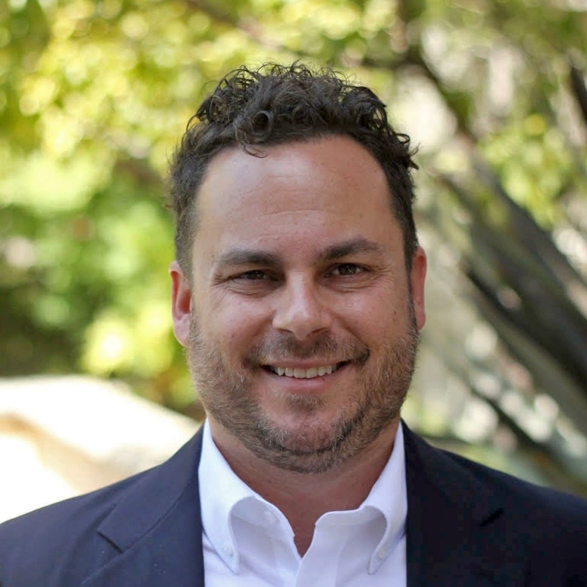 A man with short curly hair, wearing a white shirt and dark blazer, smiles outdoors. The background is blurred with green foliage and sunlight.
