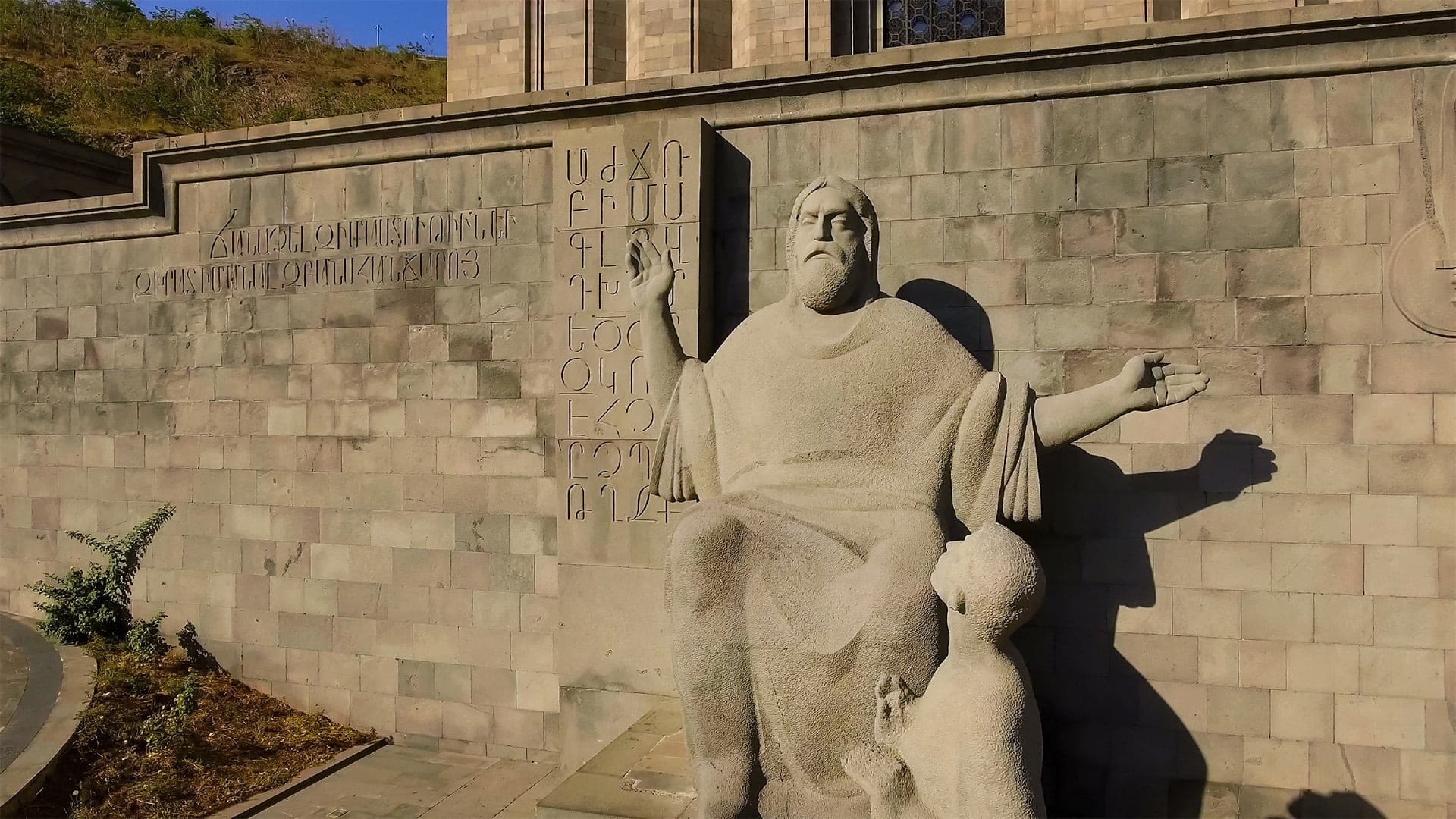 A stone statue of a seated man with a child at his feet is in front of a wall engraved with Armenian script and the Armenian alphabet, outside a gray stone building with stairs and grass nearby.