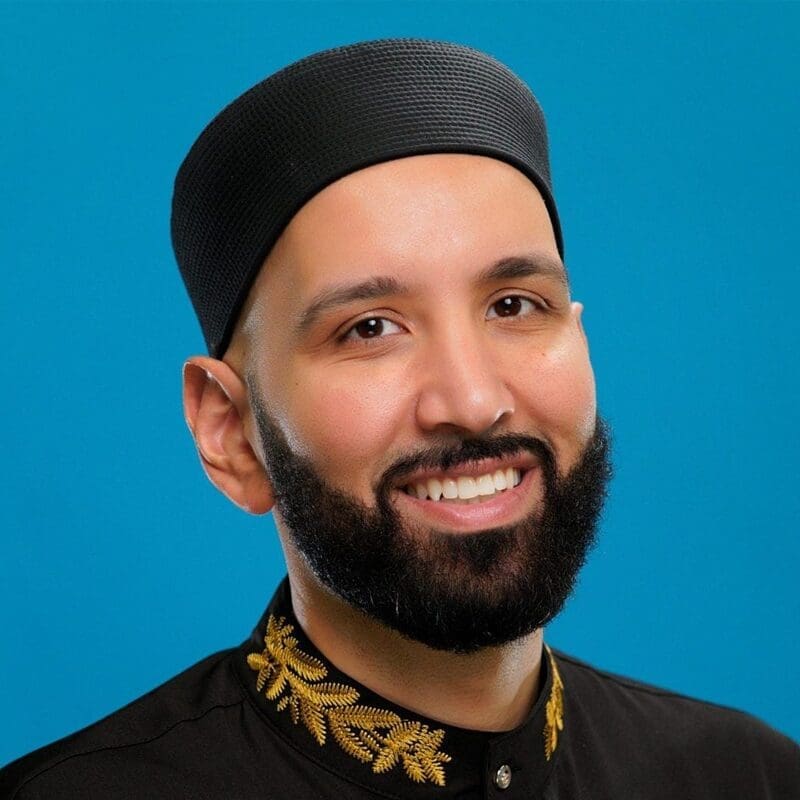 A smiling man with a dark beard wears a black hat and a black shirt with gold embroidery on the collar, posed against a bright blue background.