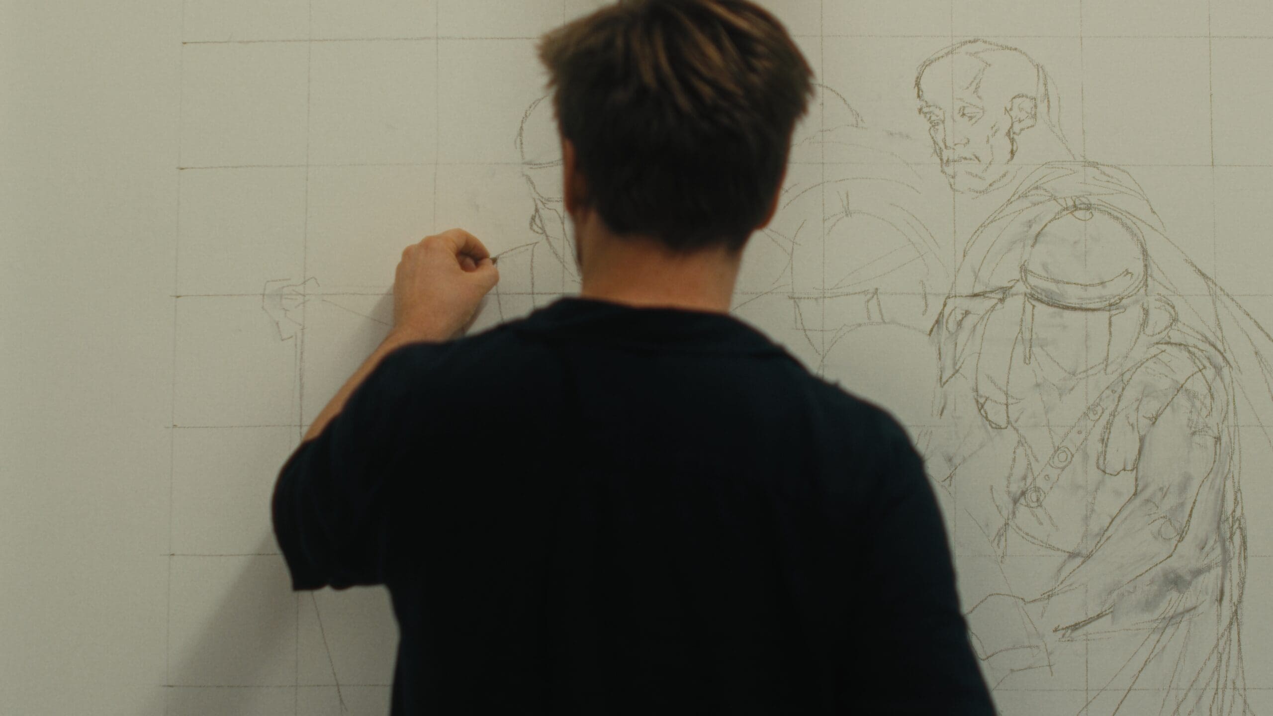 A person with short brown hair draws a detailed sketch of several people on a large white grid paper taped to the wall, seen from behind.