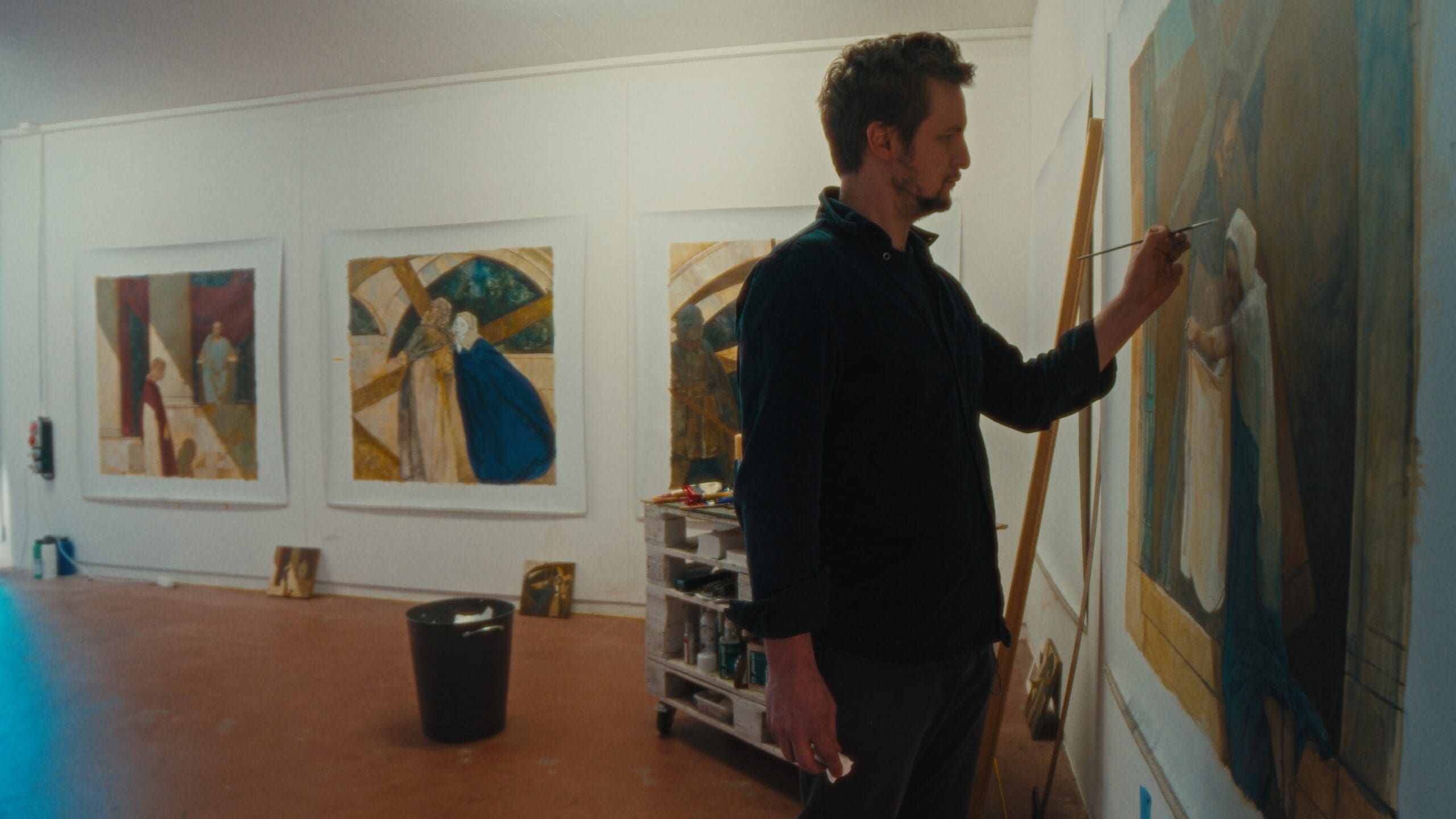 A man stands in an art studio painting a large canvas on the wall. Behind him, three colorful paintings hang, and art supplies fill a cart nearby. The room has white walls and a reddish floor.