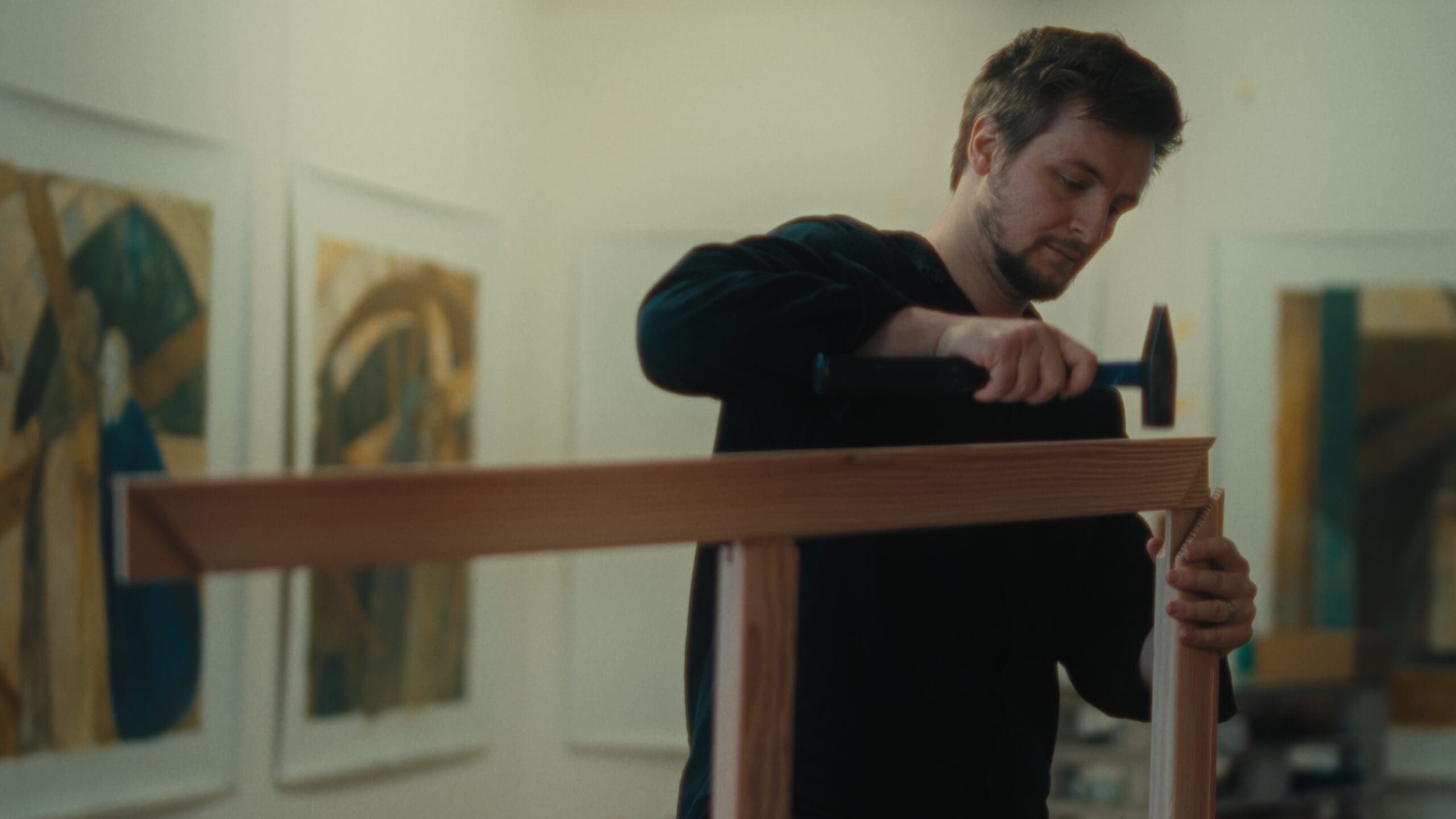 A man in a dark shirt uses a hammer to build or repair a wooden structure indoors, with abstract art hanging on the walls in the background.