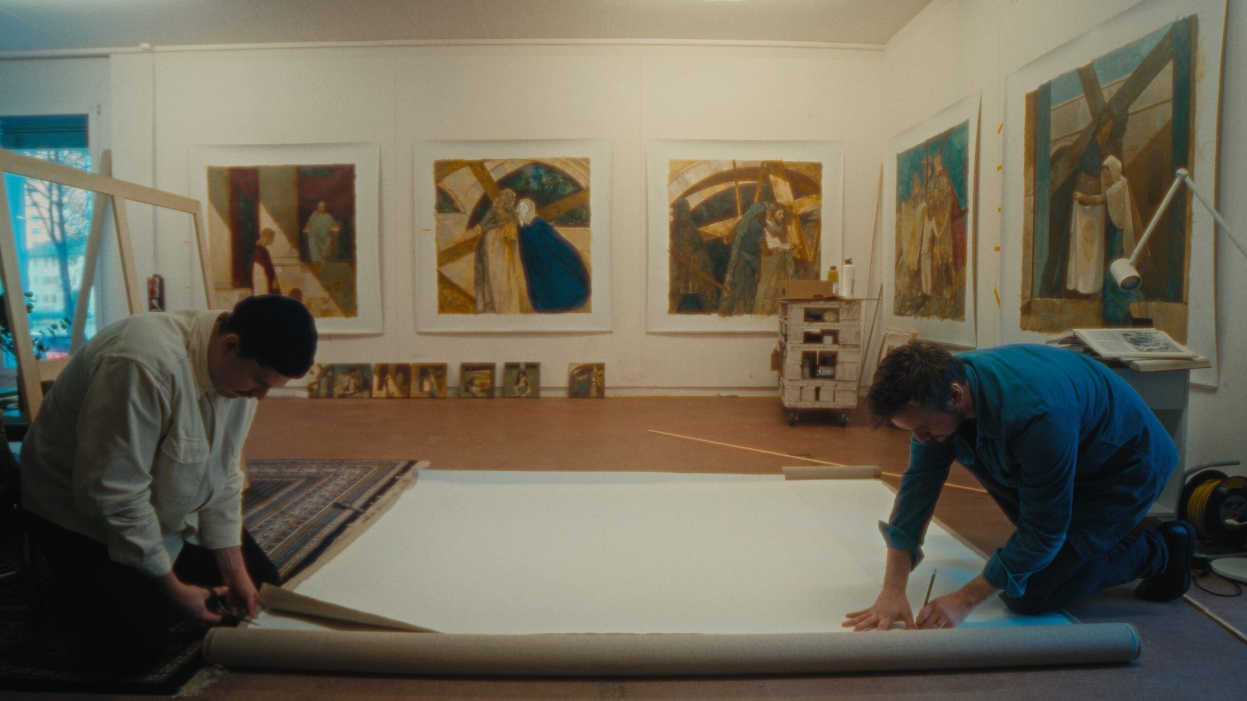 Two people unroll a large canvas on the floor of an art studio, which has several colorful, abstract paintings displayed on the walls behind them. Art supplies and tools are scattered around the room.