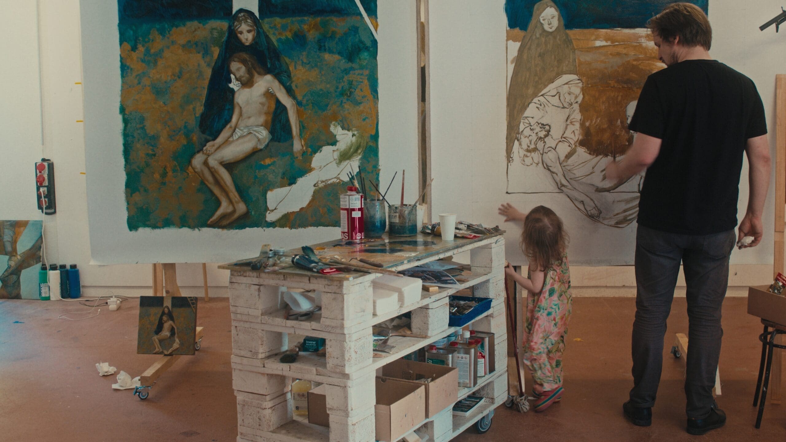 A man and a young girl stand in an art studio, painting large canvases depicting religious scenes. Art supplies and brushes are scattered on a wooden cart in the center of the room.
