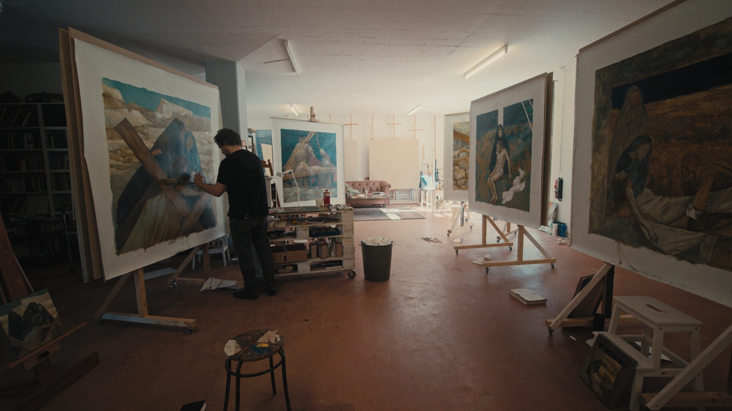 An artist stands in a studio painting a large canvas on an easel, surrounded by other finished or in-progress paintings. The room is well-lit and contains art supplies, stools, and framed artworks.