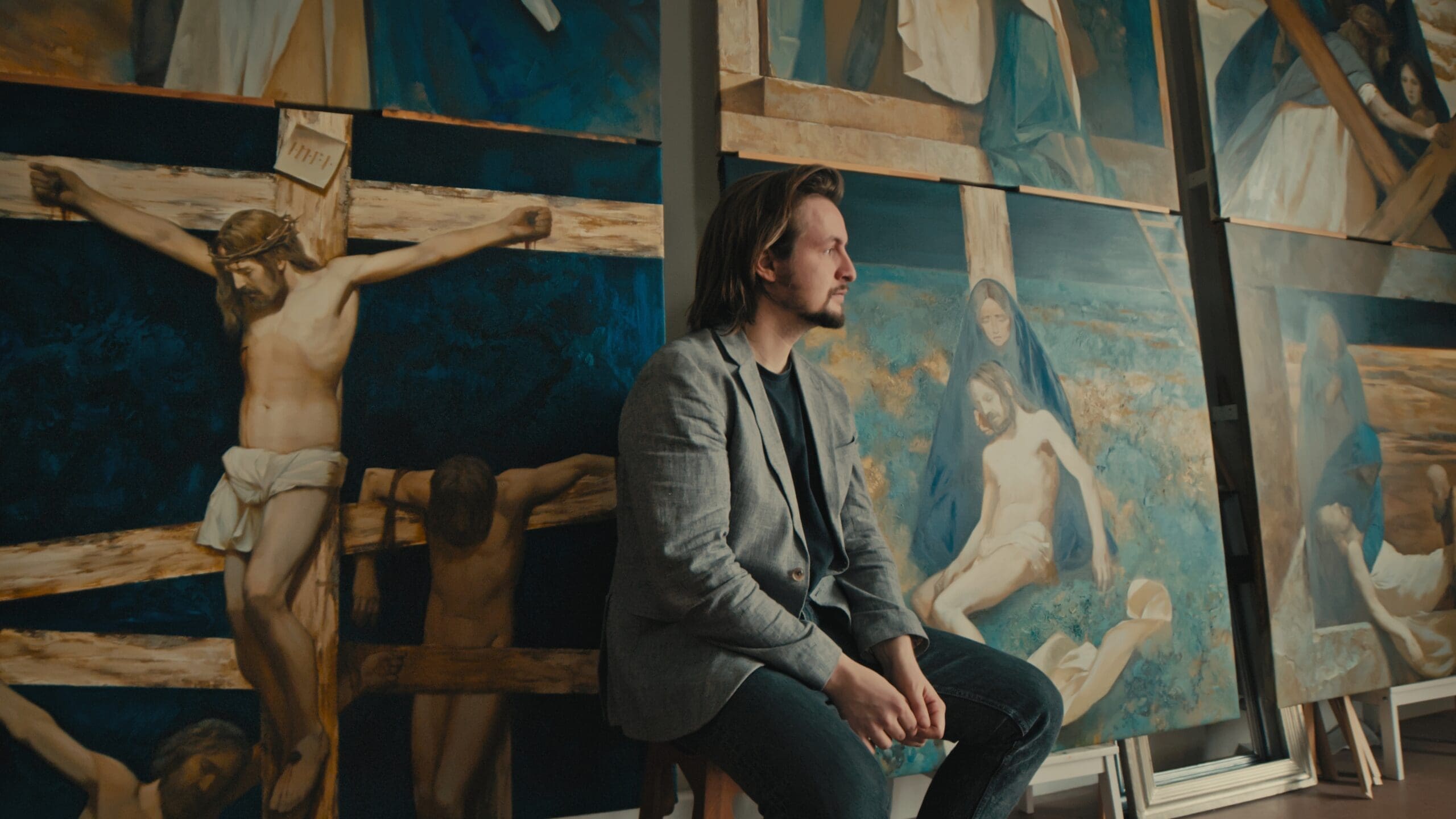A man with shoulder-length hair and a beard sits on a stool, surrounded by large paintings depicting scenes of the crucifixion of Jesus in a studio or gallery setting.