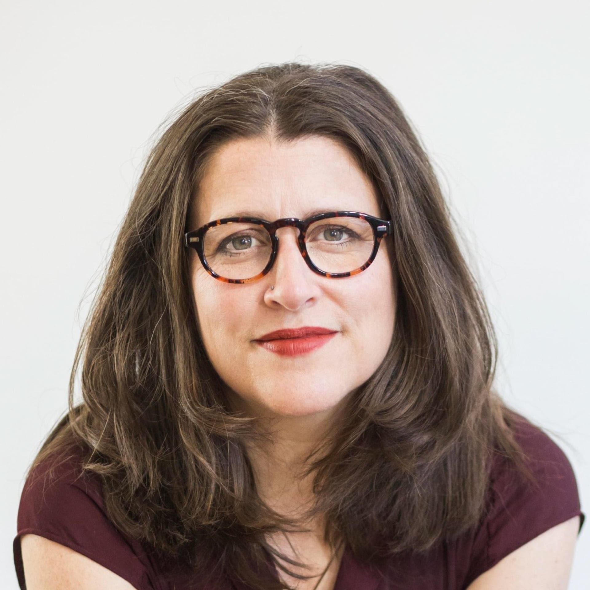 A woman with long brown hair, wearing round tortoiseshell glasses and a maroon top, looks directly at the camera with a slight smile against a plain light background.
