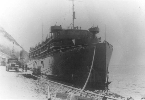 A large ship is docked at a rocky shoreline, with several people and vehicles nearby. The ship appears to be from the early 20th century, with multiple decks and antennas on top. The scene is in black and white.
