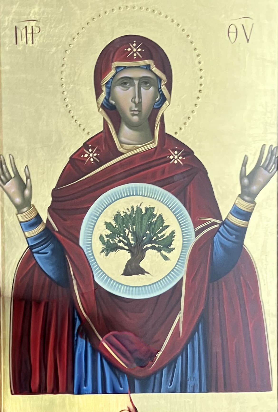 Religious icon of a woman in red and blue robes with a halo, raising her hands. A tree is depicted inside a circle on her chest. Greek letters are visible in the upper corners. Gold background.