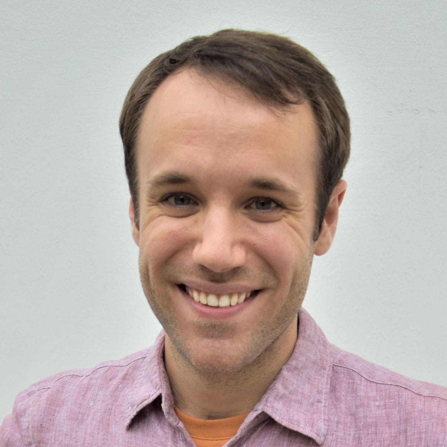 A man with short brown hair and a light beard is smiling at the camera. He is wearing a light pink collared shirt over an orange t-shirt, with a plain light background behind him.