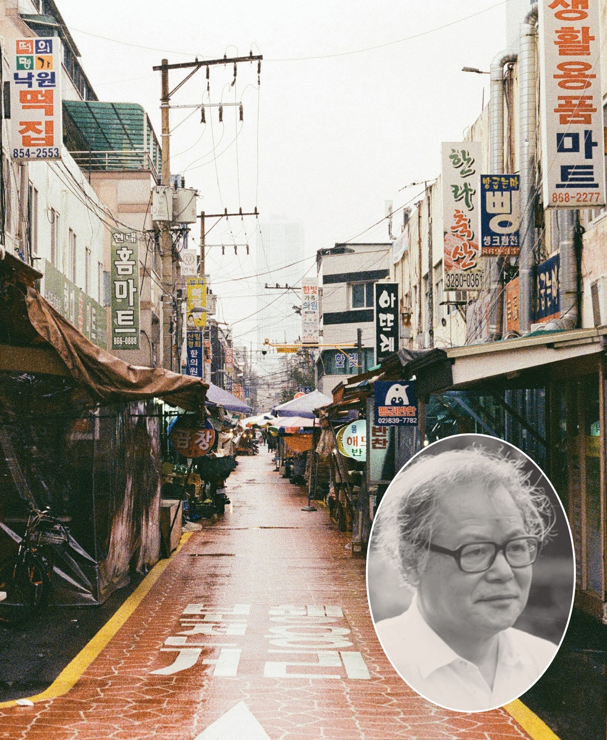 A narrow street in Korea lined with shops and colorful signs on a rainy day; an oval inset at the bottom right shows a grayscale portrait of an older man with glasses.