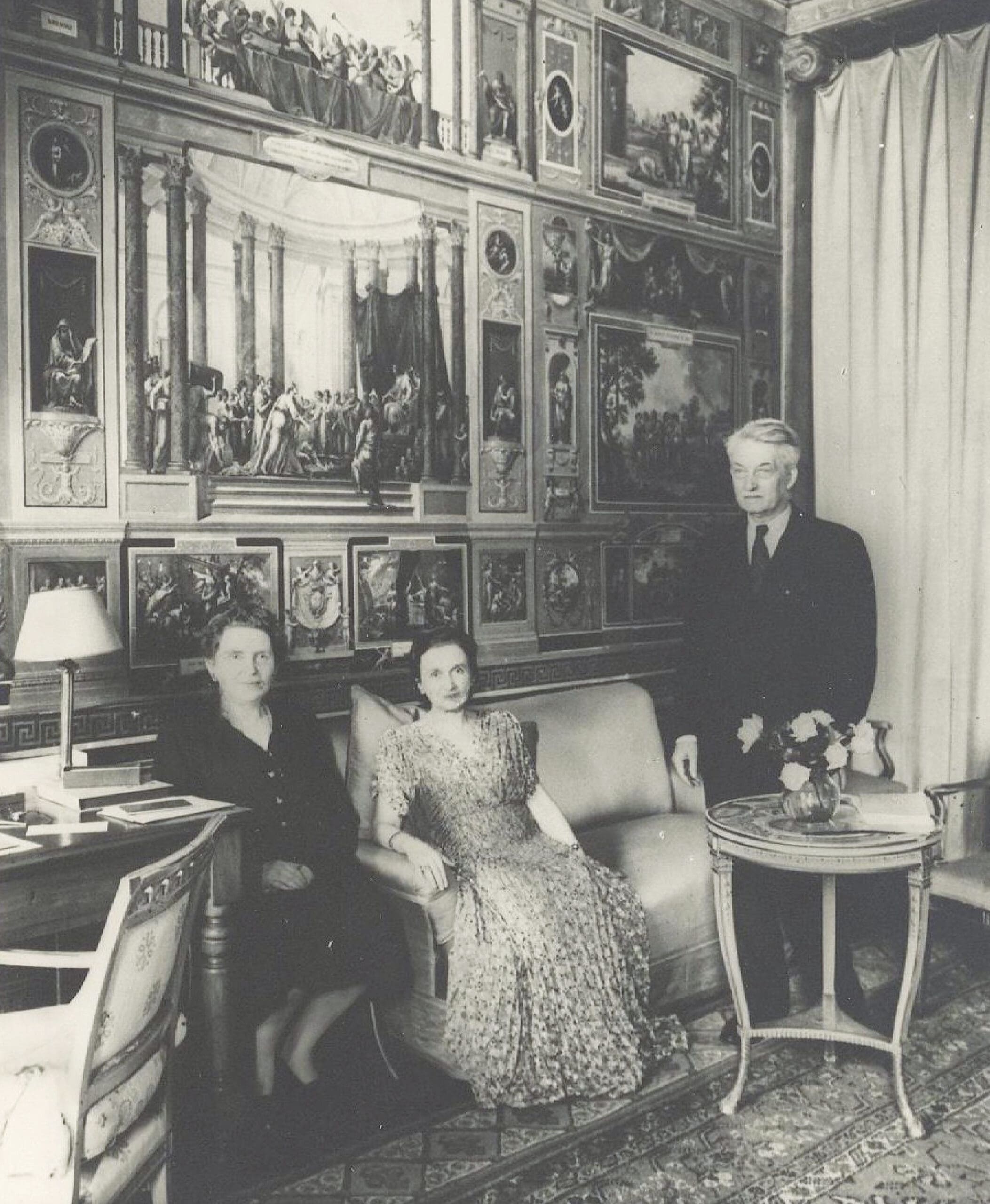 Three adults pose in an ornate room with detailed wall art and classical paintings. Two women sit on a sofa while a man stands beside a small round table with a flower vase. The room has elegant furniture and decorative patterns.