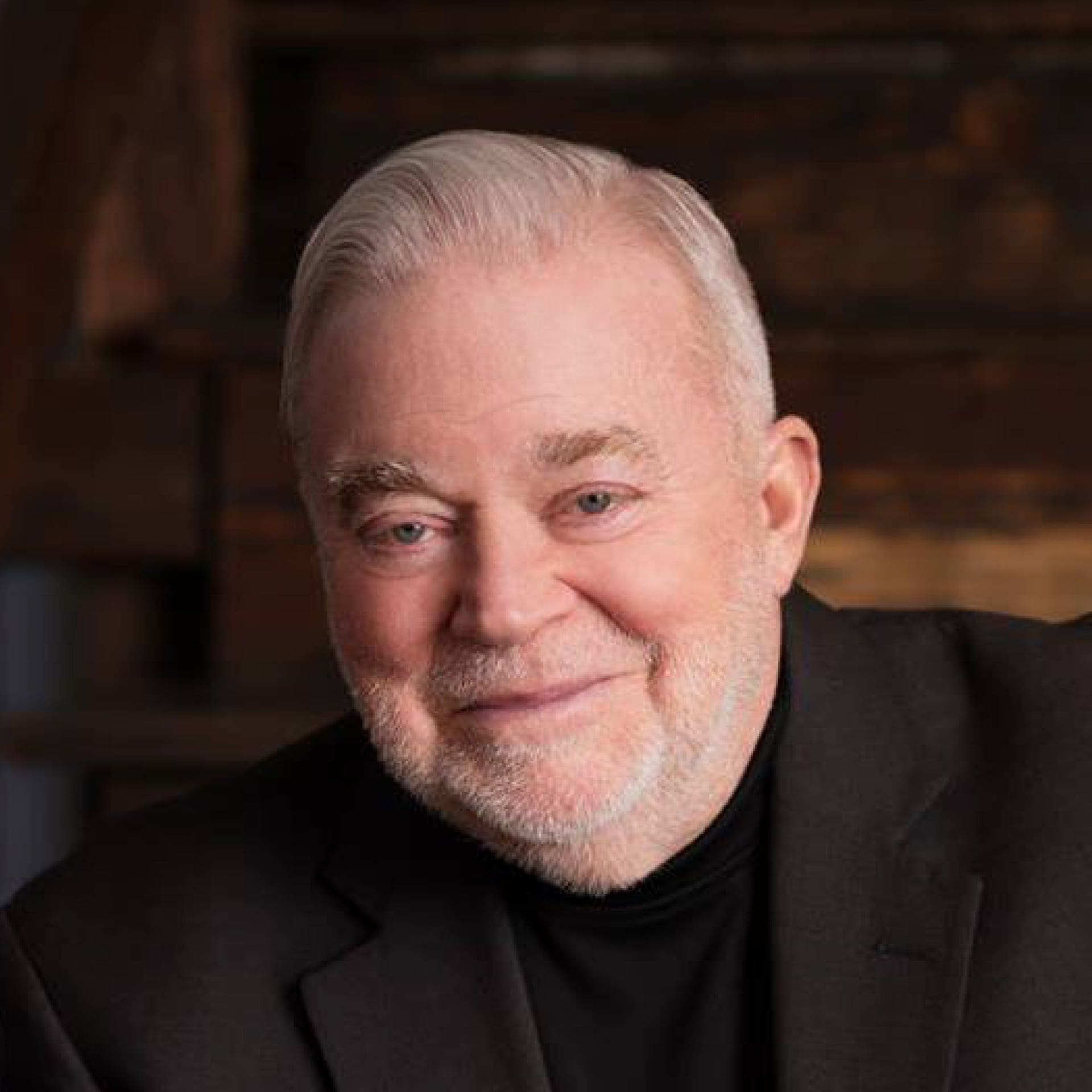 An older man with short white hair and a trimmed white beard, wearing a black suit jacket and black turtleneck, smiles softly in front of a dark, wooden background.