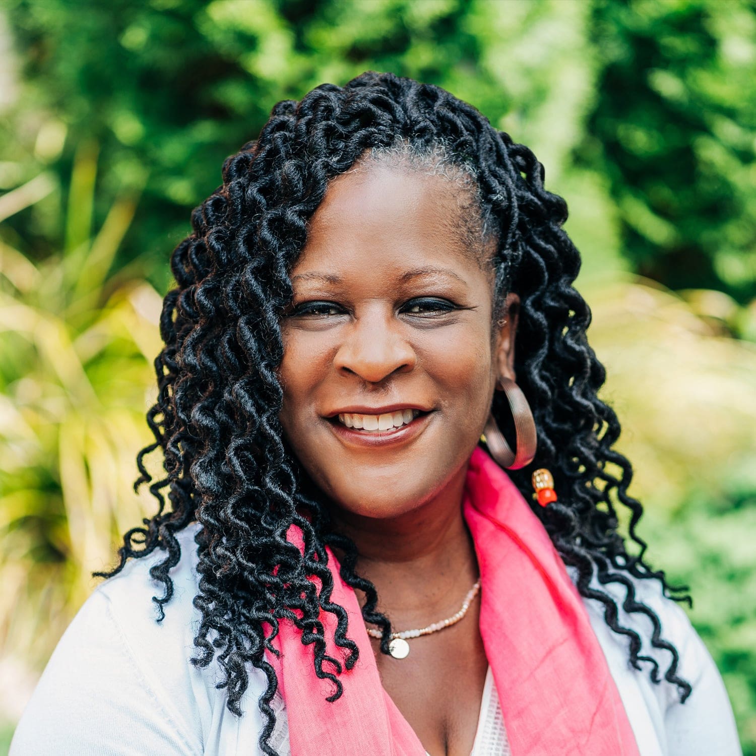 A smiling woman with curly black hair, wearing a white top, pink scarf, gold necklace, and hoop earrings, stands outdoors with greenery in the background.