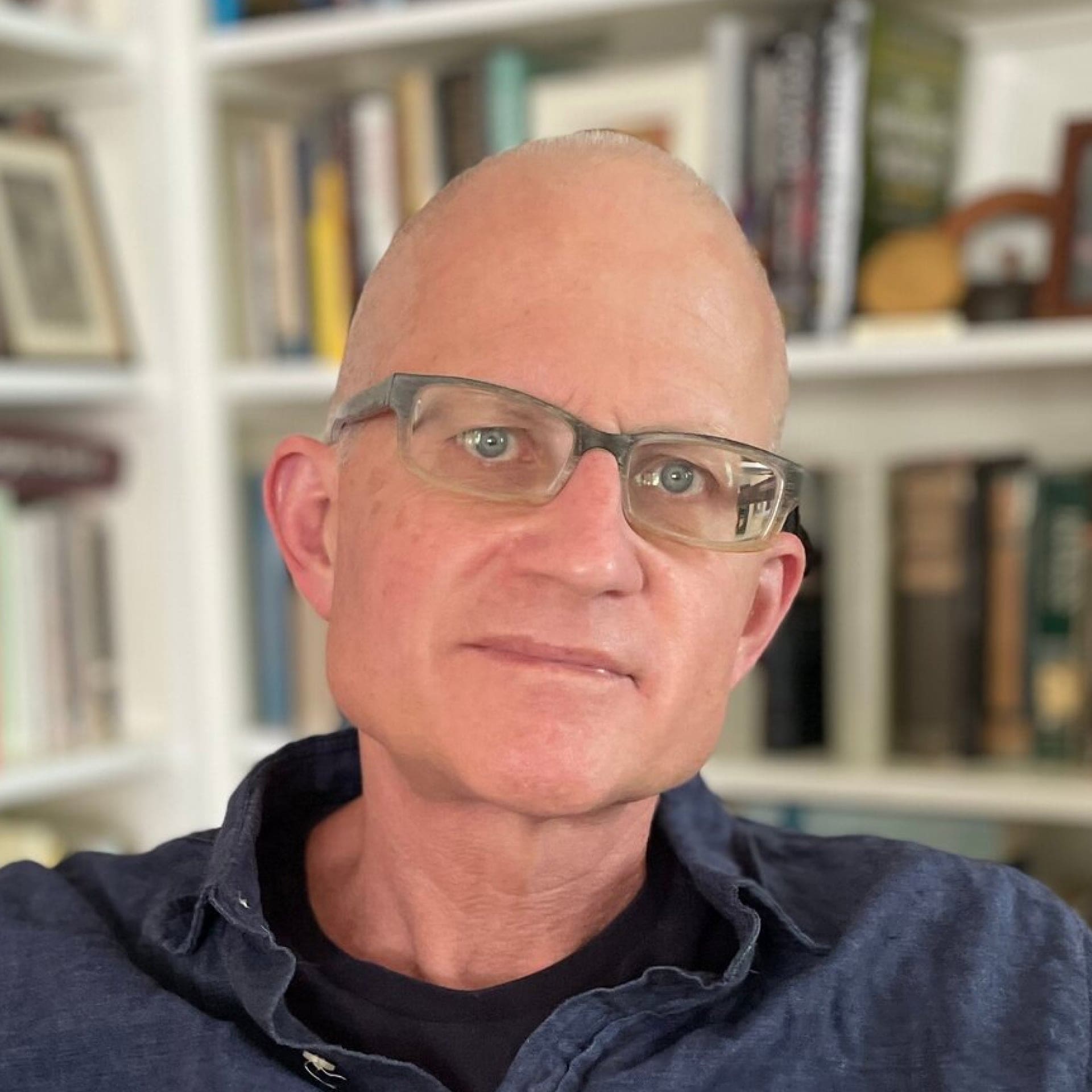 A middle-aged man with short, light hair and glasses is wearing a dark shirt and sitting in front of a bookshelf filled with books and framed photos.