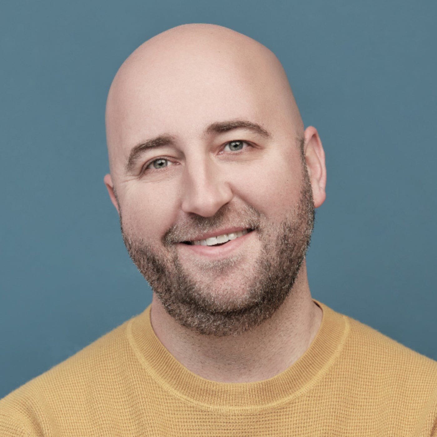 A smiling bald man with a trimmed beard, wearing a mustard yellow sweater, poses in front of a plain blue background.