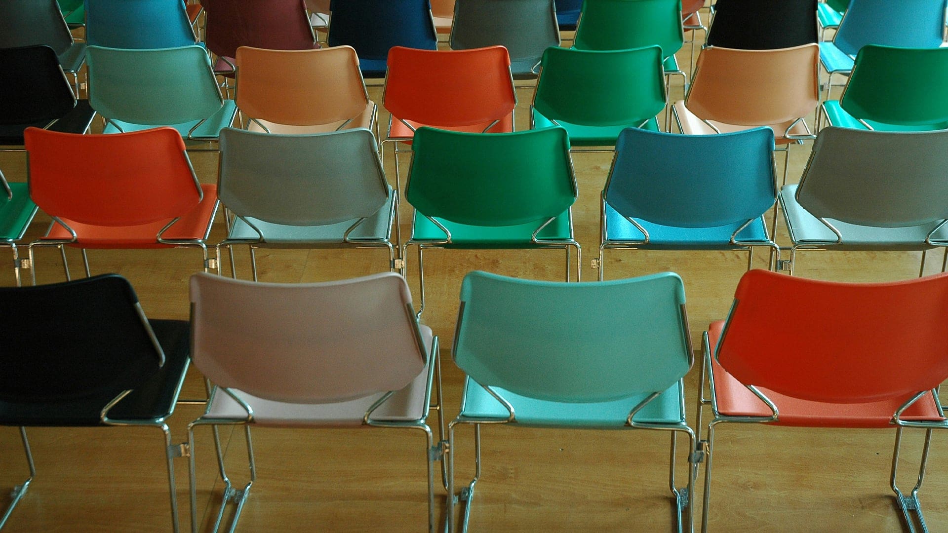 Rows of empty, colorful plastic chairs with metal legs are arranged neatly on a wooden floor, viewed from behind. The chairs are in shades of black, gray, peach, green, blue, and red.