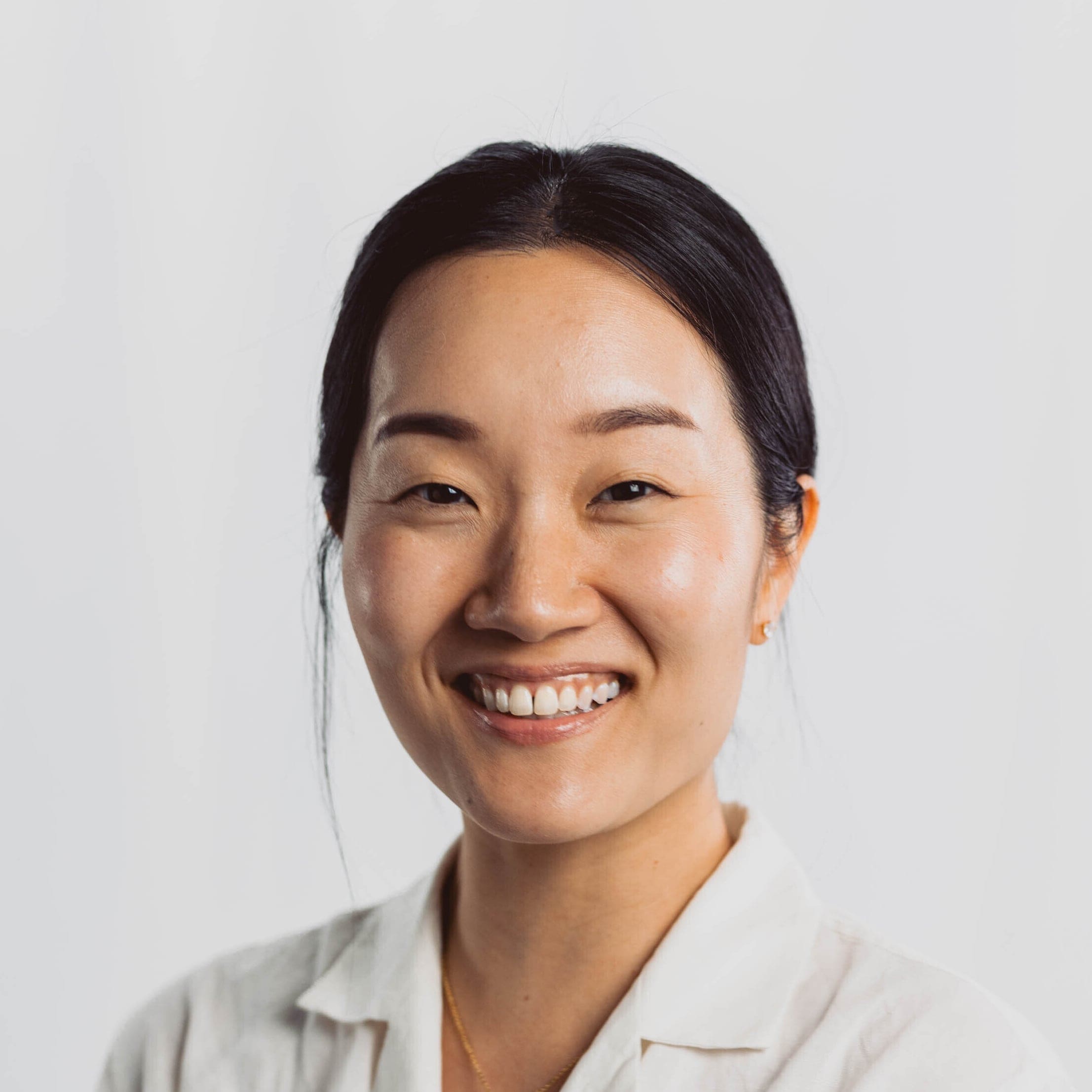 A woman with dark hair pulled back, wearing a white shirt, smiles at the camera against a plain white background.
